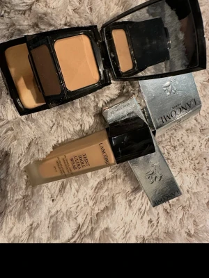 Lancome foundation och puder - Säljer Lancome Teint Idole Ultra Wear foundation i glasflaska med svart lock och tillhörande silvrig kartong, samt två Lancome puder i svarta dosor med spegel. Båda produkterna har en beige nyans som passar till en naturlig look.
