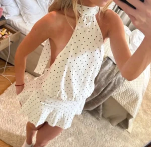 Vit prickig kortklänning från Jacq & Leon - Supersöt vit kortklänning med svarta prickar från Jacq & Leon. Klänningen har omlott-design, hög hals med knyt och volangdetaljer vid nederkanten. Ärmlös och med öppen rygg, perfekt för sommarens alla tillfällen. Materialet känns mjukt och lätt.