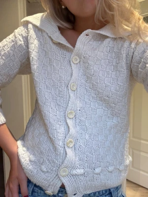 Vit stickad kofta med stora knappar - Säljer en vit stickad kofta med snyggt rutmönster och stora, ljusa knappar framtill. Koftan har en bred krage och trekvartsärmar, samt en dekorativ stickad detalj längst ner. Perfekt att slänga på över en t-shirt eller linne.