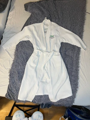 Vit badrock från Studentbostäder i Norden AB - Vit badrock med broderad logga från Studentbostäder i Norden AB. Rocken har två stora fickor framtill och ett knytbälte i midjan. Materialet är bomull med en våfflad struktur som känns mjuk mot huden. Perfekt att slänga på sig efter duschen eller när du chillar hemma.