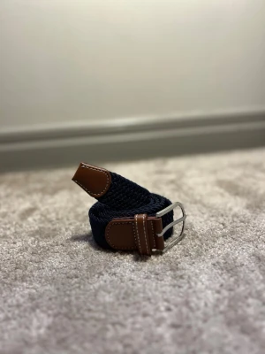 Flätat bälte marinblå  - Ett stilrent flätat bälte från Ragazzo i en klassisk marinblå färg. Bältet är 105 cm långt och tillverkat i slitstarkt material som ger både komfort och hållbarhet. Den flätade designen gör bältet lätt att justera för optimal passform och passar lika bra till vardag som till mer uppklädda tillfällen.