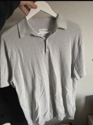 Ljusgrå polotopp från These Glory Days - Säljer en ljusgrå polotopp från These Glory Days i silk blend-material. Toppen har klassisk krage, korta ärmar och två knappar framtill. Perfekt för dig som gillar stilrena och enkla plagg med mjuk känsla.