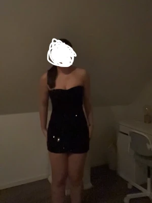 Svart glittrig axelbandslös klänning - Säljer en svart, glittrig kortklänning med axelbandslös design. Klänningen har en tight passform. Materialet är elastiskt med paljetter på. Supersnygg till fest!