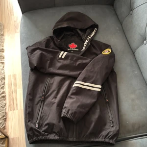 Svart vindjacka från North Canada - Svart vindjacka med huva och dragkedja från North Canada. Jackan har två vita ränder på ena ärmen, broderad patch på axeln och två framfickor med dragkedja. Perfekt för blåsiga dagar.