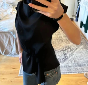 Svart blus - Säljer en svart blus från Zara med snygg omlott-detalj och knyt i midjan. Blusen har korta ärmar och en draperad halsringning som ger en cool och elegant look. Materialet är mjukt och har en lätt glansig finish som känns lyxig.