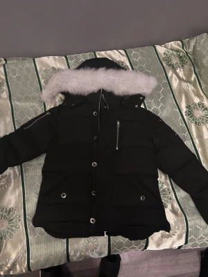 Svart Moose Knuckles dunjacka med päls - Säljer en svart dunjacka från Moose Knuckles med vit päls på huvan. Jackan har flera fickor med dragkedja och knappar, samt ett snyggt logomärke på ärmen. Perfekt för kalla dagar med sin tjocka och quiltade design.