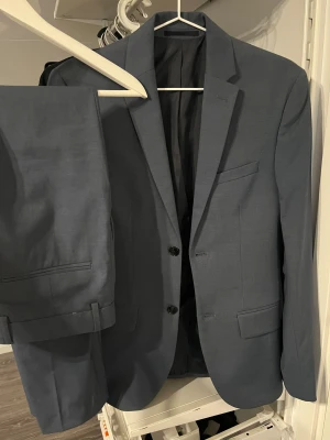 Blå kostym från H&M - Säljer en stilren blå kostym med matchande kavaj och kostymbyxor. Byxorna är raka och har bälteshällor. Materialet är främst polyester. Kostymen använde jag en gång och är i storleken S. Nypris: 1150kr