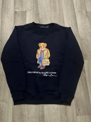 Svart sweatshirt Polo Bear Ralph Lauren - Svart sweatshirt från Polo Ralph Lauren i storlek S med ikoniskt Polo Bear-tryck på framsidan. Rund hals, ribbade muddar och mjukt bomullsmaterial. Perfekt för dig som gillar streetstyle och vill ha en klassisk look med en twist.