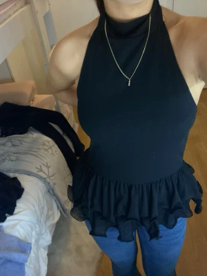 Svart volangtop med hög hals - Säljer en svart topp med hög hals och ärmlös design. Toppen har en snygg volangkant nedtill som ger en cool och feminin touch. Materialet känns mjukt och lätt, perfekt att styla med jeans eller kjol. Passformen är normal och toppen har en stilren look.