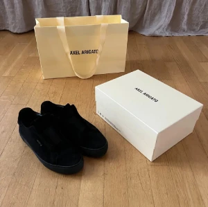 Svarta Axel Arigato Clean 360 sneakers - Säljer ett par svarta Axel Arigato Clean 360 Laceless sneakers i mocka. Skorna har minimalistisk design utan snörning, med elastisk panel över vristen och diskret logga i guld på sidan. Snygg rund tå och platt sula, perfekt för dig som gillar stilrena och moderna sneakers.