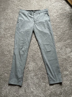 Grå kostymbyxor från J.Lindeberg - Snygga grå kostymbyxor från J.Lindeberg med klassisk rak passform och diskreta fickor med knapp bak. Byxorna har bälteshällor och är gjorda i ett mjukt material som känns lyxigt. Perfekta för dig som gillar stilrena och moderna plagg.