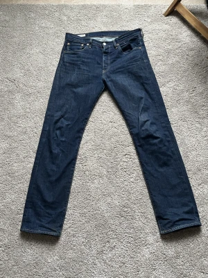 Levi's 501 mörkblå jeans W31 L32 straight fit - Säljer ett par klassiska Levi's 501 jeans i mörkblå tvätt. De har rak passform, fem fickor och snygga kontrastsömmar. Ikonisk läderpatch bak i midjan och röd Levi's-tag på bakfickan. Perfekt för dig som gillar tidlös stil och jeans av hög kvalitet.
