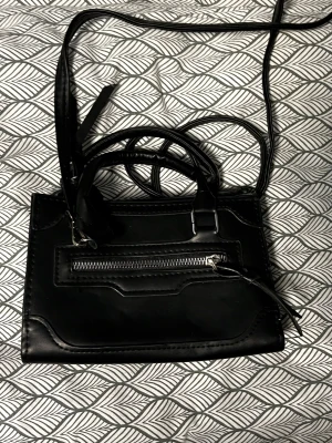 Svart axelväska med dragkedja - Snygg svart axelväska i skinnimitation med både handtag och lång axelrem. Väskan har en praktisk dragkedja framtill och stilrena detaljer. Perfekt storlek för det viktigaste och passar till många olika outfits.