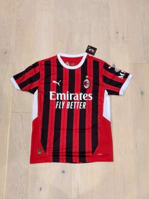 Ac Milan football Jersey  - Bilderna visar en AC Milan fotbollströja från Puma i röd och svart med vita detaljer, storlek M. Ingen sko finns med på bilderna.