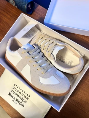 Maison Margiela sneakers beige/vit - Maison Margiela sneakers i beige och vitt med klassisk gummisula. Ovandel i skinn och mocka, snygga kontraster mellan material och färger. Låg modell med rund tå och snörning. Perfekta för dig som gillar stilrena och trendiga sneakers.