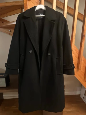 Svart kappa från Zara i ullmix - Säljer en svart kappa från Zara i storlek S. Klassisk modell med bred krage, dubbelknäppning och fickor fram. Kappan har långa ärmar med knappdetalj vid mudden och är tillverkad i en mjuk ullmix. Perfekt för höst och vinter. Tyvärr har insidan av ärmarna börjat bli lite noppiga men det går ju att ta bort!