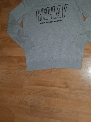 Grå sweatshirt från Replay - Säljer en grå sweatshirt från Replay med stort svart tryck på bröstet där det står 'REPLAY DENIM GOODS SINCE 1981'. Tröjan har rund hals, långa ärmar och ribbade muddar vid ärmslut och nederkant. Perfekt för chill dagar och enkel att matcha med jeans.