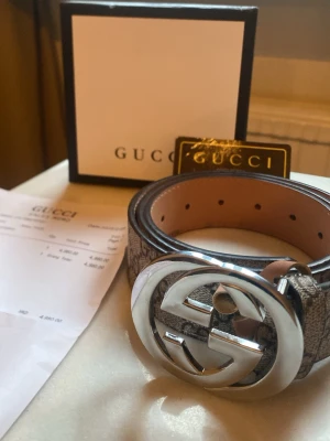 Gucci bälte - Säljer ett snyggt Gucci-bälte i beige med klassiskt GG-mönster och ett stort, silverfärgat GG-spänne. Bältet är gjort i äkta skinn och har detaljer i mörkbrunt på insidan. Perfekt accessoar för att lyfta din outfit med lyxig känsla.
