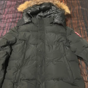 Svart dunjacka från Canada Goose - Säljer en svart dunjacka från Canada Goose med stor huva och snygg pälsdetalj. Jackan har ett klassiskt märke på ärmen, knappar framtill och quiltad design. Perfekt för kalla dagar och har en riktigt schysst look.