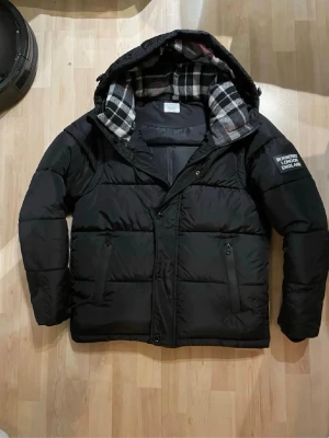 Svart pufferjacka från Burberry - Svart pufferjacka från Burberry med avtagbara ärmar så den kan användas som väst. Jackan har huva med rutigt foder i svart, vitt och rött, samt patch med Burberry London England på ärmen. Flera fickor med dragkedja och knappstängning framtill.