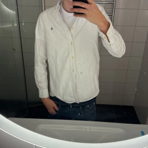 Vit skjorta från Ralph Lauren - Säljer en klassisk vit skjorta från Ralph Lauren med knappar framtill och lång ärm. Skjortan har en liten broderad logga på bröstet och är gjord i mjuk bomull. Perfekt till jeans eller chinos för en clean och stilren look. Lite liten på mig , passar någon som är 180 och väger 65.