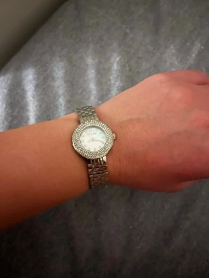 Silverfärgad klocka Ivy N Rox - Snygg och stilren klocka från Ivy N Rox i silverfärg med rund urtavla och glittrande stenar runt boetten. Urtavlan är vit med små kristaller som timmarkeringar och romersk tolvmarkering. Länken är klassisk och metallisk, perfekt för att lyfta din outfit.