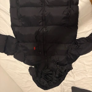 Svart pufferjacka från Polo Ralph Lauren - Säljer en svart pufferjacka från Polo Ralph Lauren med huva och dragkedja framtill. Jackan har två bröstfickor med dragkedja och den klassiska röda Polo-loggan på bröstet. Perfekt för kalla dagar och riktigt snygg till streetwear-stilen.