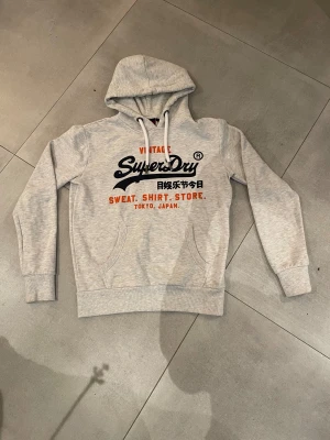 Superdry hoodie  - Tja! Säljer nu en riktigt snygg grå hoodie från Superdry med ett snyggt tryck framtill med knappast några defekter alls bara ett minimalt som inte syns på sista bilden så skicket är 9,5/10. Storlek M och passar killen på bilden som är 185! Hör av dig vid minsta lilla fundering eller fråga!🤗mvh EM_resell