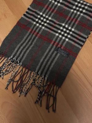 Rutig grå halsduk från Burberrys - Säljer en klassisk rutig halsduk från Burberrys i 100% lammull. Den har en snygg grå bas med inslag av vitt, svart och rött i mönstret. Halsduken har fransar i ändarna och en vävd Burberrys-etikett. Perfekt för att ge din outfit en tidlös touch.