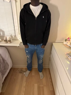 Polo Ralph Lauren svart zip hoodie – stl M - Svart zip hoodie från Polo Ralph Lauren i storlek M. Klassisk modell med huva, dragkedja framtill och den ikoniska vita Polo-loggan broderad på bröstet. Mjuk och bekväm kvalitet, perfekt för vardagsbruk.