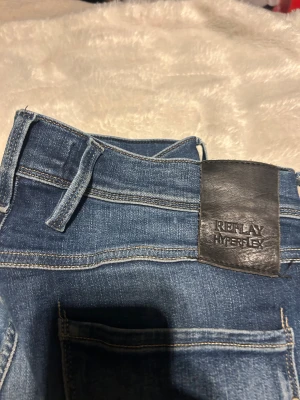Replay Hyperflex blå jeans - Säljer ett par Replay Hyperflex jeans i klassisk blå färg med snygga slitningar och kontrastsömmar. Jeansen har fem fickor, bälteshällor och en bekväm passform. Ett litet hål som man knappt märker mellan benen. Storlek 32 32