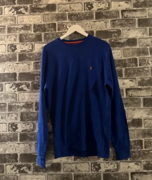 Blå långärmad tröja från Polo Ralph Lauren - Blå långärmad tröja från Polo Ralph Lauren med orange logobrodyr på bröstet och en orange detalj vid halsringningen. Tillverkad i 100% bomull, perfekt för dig som gillar klassisk stil med en färgklick. Enkel och snygg design som funkar året runt.