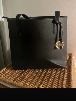 Svart axelväska från Anna Field - Snygg svart axelväska från Anna Field i stilren design med rektangulär form. Väskan har långa axelband och ett dekorativt guldfärgat hänge med logga. Materialet är slätt syntet och insidan har en praktisk ficka med dragkedja.