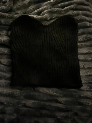 Svart ribbad stickad tubtopp - Säljer en svart ribbad tubtopp i stickat material. Toppen har en bandeauformad överdel och är helt ärmlös, perfekt för att styla med höga jeans eller kjol. Enkel och stilren design som passar till många olika looks.