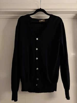 Svart kofta från Ströms Stockholm - Snygg svart kofta från Ströms Stockholm med klassisk v-ringning och sex silverfärgade knappar framtill. Koftan har långärmade och ribbade muddar vid ärmslut och nederkant. Perfekt för lager-på-lager och enkel att matcha med olika stilar. Ny pris 1799kr 