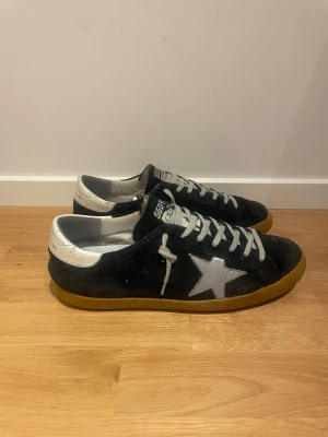 Golden Goose  - Golden Goose sneakers i svart mocka med vit stjärndetalj på sidan och vit häl med logga. Skorna har snörning och en brun gummisula med svart undersida. Klassisk rund tå och platt sula, perfekt för dig som gillar streetstyle.