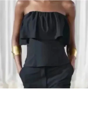 SÖKER 123 - Snygg svart tubtopp med volang upptill som ger en cool och trendig look. Toppen är axelbandslös och har en bandeau-stil som passar perfekt till sommarens outfits. Materialet känns lätt och bekvämt mot huden.