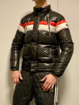 Svart pufferjacka från Hugo Boss - Säljer en svart pufferjacka från Hugo Boss med feta röda och vita ränder över bröst och rygg. Jackan har hög krage, dragkedja och två zip-fickor framtill. Insidan har Boss-logga i mönstret. Perfekt för kalla dagar och riktigt snygg streetstil. Storlek S eller liten M. Personen på bilden är 180 cm.