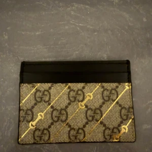 Gucci GG Supreme plånbok i skinn - Säljer en snygg och stilren plånbok från Gucci med klassiskt GG Supreme-mönster i beige och svart, dekorerad med diagonala guldiga linjer. Plånboken är tillverkad i skinn och har flera fack för kort. Perfekt för dig som gillar lyxiga accessoarer.