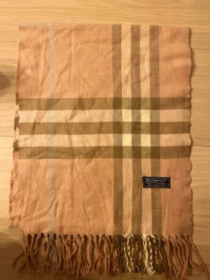 Rutig beige kashmirväst från Burberrys - Säljer en klassisk rutig väst från Burberrys i mjuk 100% kashmir. Västen har ett ikoniskt beige och brunt rutmönster med fransar nedtill. Perfekt för dig som vill ha en tidlös och lyxig look med brittisk touch.