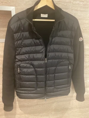 Svart cardigan från Moncler - Snygg svart cardigan från Moncler med quiltad front och ärmar i stickad kvalitet. Jackan har dragkedja framtill och två fickor med dragkedja. Klassisk Moncler-logga på ena ärmen och hög krage för extra stilpoäng.