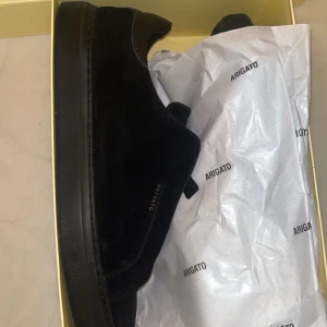 Svarta sneakers från Axel Arigato - Snygga svarta sneakers från Axel Arigato med stilren design. Skorna har en platt sula, rund tå och är tillverkade i mocka med detaljer i skinn. Diskret Arigato-logga i guld på sidan. Perfekta för dig som gillar minimalistisk och modern stil.