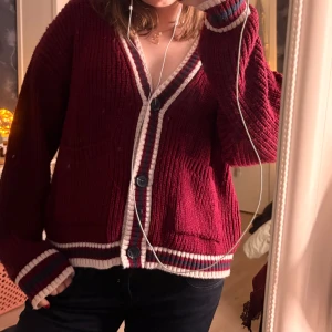 Vinröd stickad kofta med knappar - Säljer en vinröd stickad kofta med vita och mörkröda ränder längs kanterna och muddar. Koftan har v-ringad hals, stora svarta knappar och två fickor framtill.