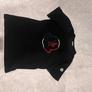 Svart Moncler t-shirt  - Svart t-shirt från Moncler med stor broderad logga i rött, blått och vitt på bröstet samt en mindre logotyp på ärmen. Klassisk rund hals och korta ärmar. Materialet är mjuk bomull som känns skön mot huden. Perfekt för dig som gillar exklusiva streetwear-plagg.
