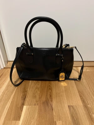 Svart handväska från Ralph Lauren - Stilren svart handväska från Ralph Lauren i skinn med både handtag och axelrem. Väskan har flera fack med dragkedja och snygga gulddetaljer. Klassisk och rymlig modell som rymmer allt du behöver. Perfekt för dig som gillar en clean och lyxig look.