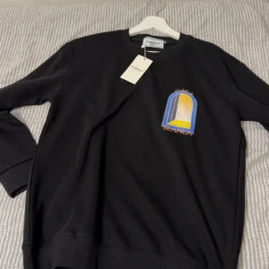 Svart sweatshirt från Casablanca - Svart sweatshirt från Casablanca med färggrafiskt tryck i blått, gult och orange både fram och bak. Tröjan har rund hals, långa ärmar och en relaxed passform. Perfekt för dig som gillar stilrena streetwear-plagg med coola detaljer.