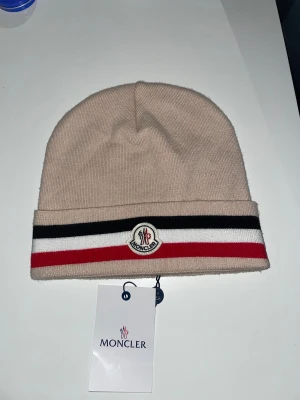 Beige Moncler mössa med ränder - Snygg beige mössa från Moncler med klassiska svarta, vita och röda ränder på uppviket. Mössan har Moncler-logga framtill och är stickad i mjukt material som passar perfekt när det är kallt ute. Stilren och enkel att matcha med din vinterjacka.