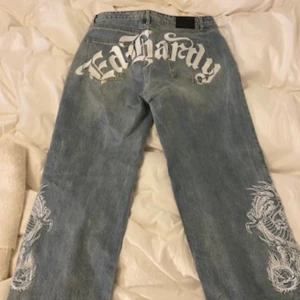 Ed Hardy x Urban Outfitters jeans  - Säljer ett par blå jeans från Ed Hardy x Urban Outfitters med stort vitt Ed Hardy-tryck över bakfickorna och detaljerade vita drakmotiv längs båda benen. Jeansen har rak passform och klassisk femficksdesign. Perfekt för dig som gillar statement-plagg.