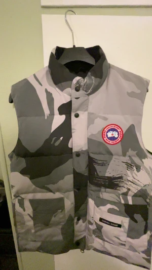 Grå camo dunväst från Canada Goose - Säljer en Canada Goose dunväst i storlek L med grått och svart camouflage-mönster. Västen har hög krage, två stora fickor framtill och klassisk Canada Goose-patch på bröstet. Perfekt för lager-på-lager och streetwear-vibe.