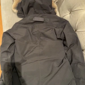 Canada Goose - Säljer en svart parkas från Canada Goose med päls på huvan och snygga detaljer som knappar och dragkedja. Jackan har lång ärm, flera fickor och klassisk logga på bröstet. Perfekt för kalla dagar med sin fodrade insida och robusta material.